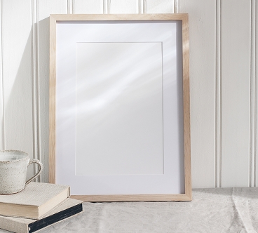 Empty wooden frame mockup on beige linen tablecloth background. White wooden wall paneling background. Artistic still life with cup of coffee and books in sunlight, Scandinavian interior. Art concept.
