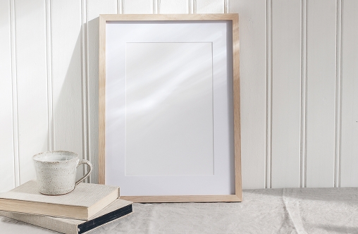Empty wooden frame mockup on beige linen tablecloth background. White wooden wall paneling background. Artistic still life with cup of coffee and books in sunlight, Scandinavian interior. Art concept.