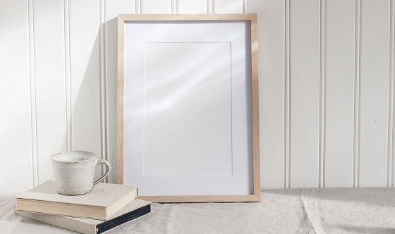 Empty wooden frame mockup on beige linen tablecloth background. White wooden wall paneling background. Artistic still life with cup of coffee and books in sunlight, Scandinavian interior. Art concept.